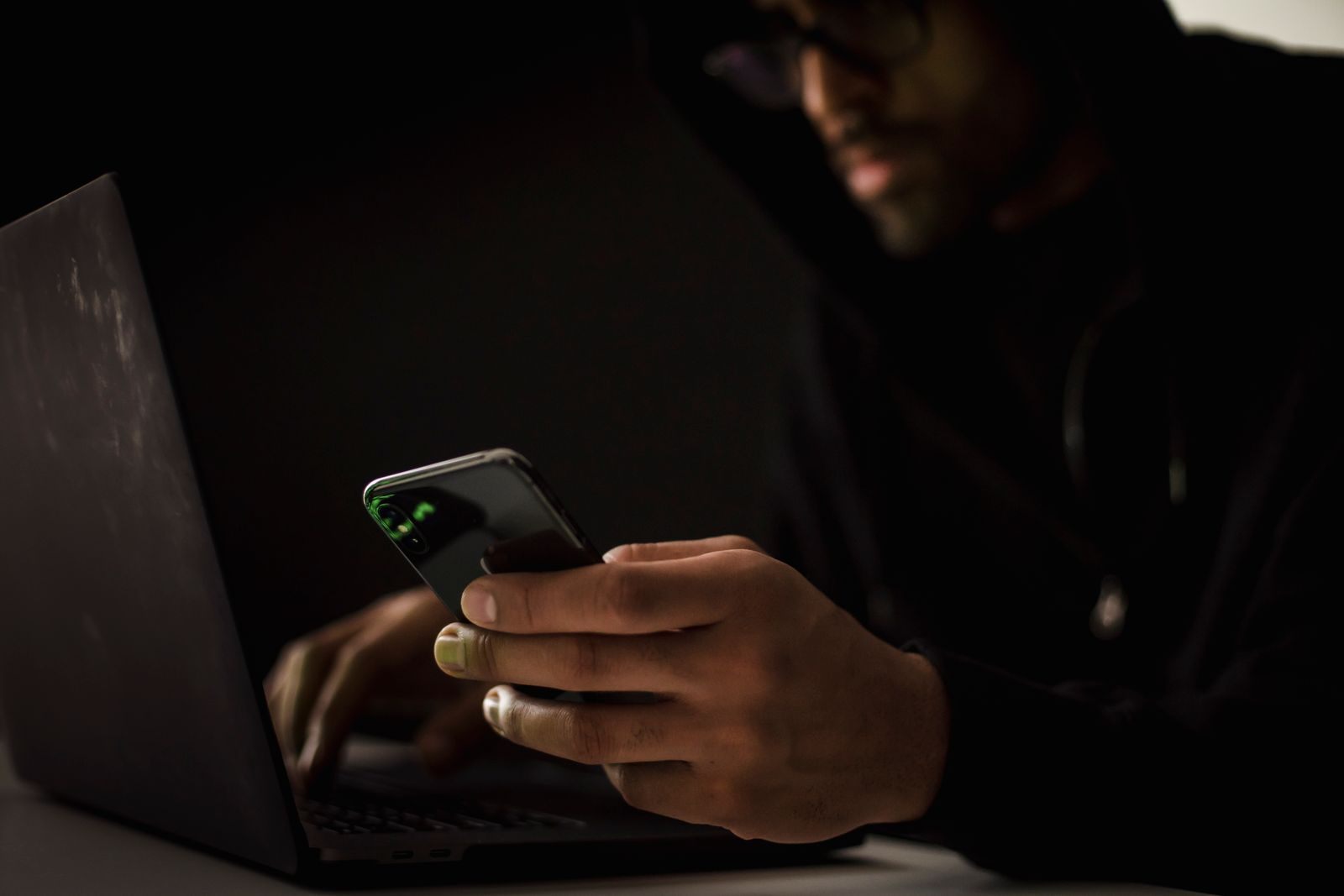 Hooded person in a dark room using a phone and a laptop